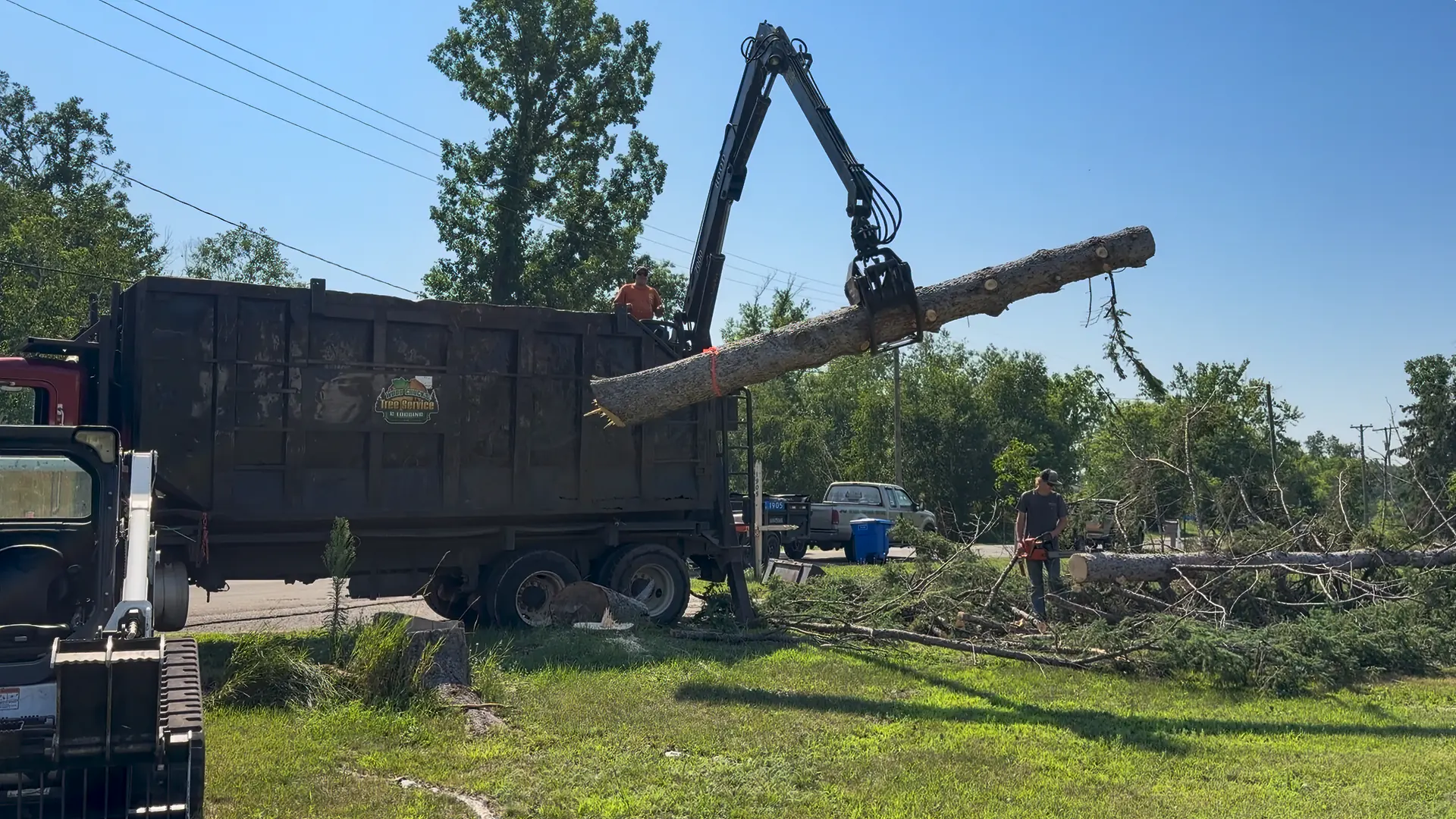 WoodChucks grapple truck lifting a sectioned tree trunk into the dump trailer while a crew member works below